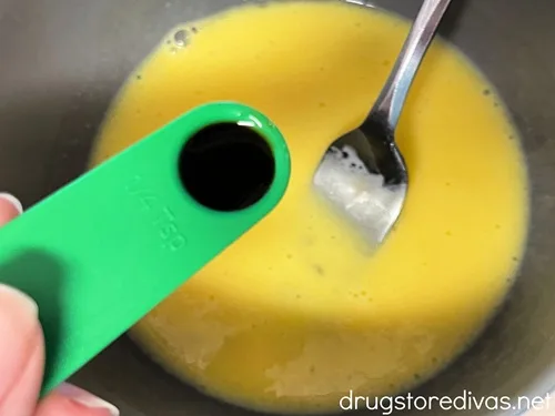 A green measuring spoon with vanilla extract it in above a bowl with scrambled raw egg with a fork in it.