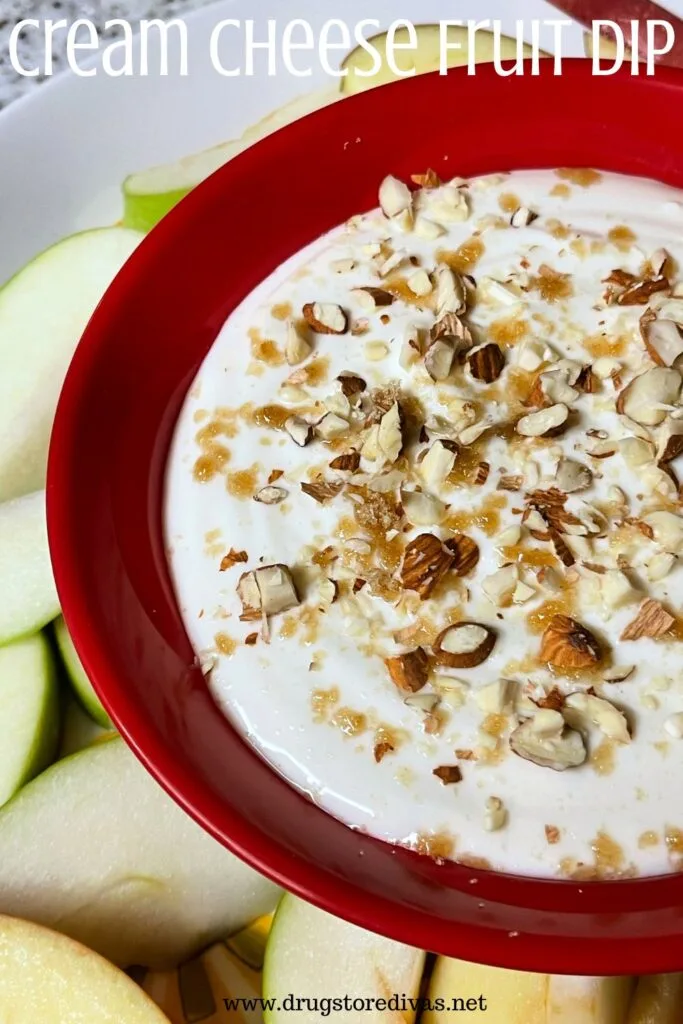 A white dip with brown sugar and nuts on top in a red bowl next to slices green apples and the words "Cream Cheese Fruit Dip" digitally written on top.