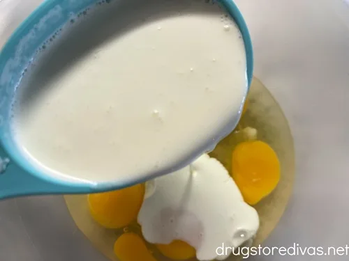 Heavy cream in a blue measuring cup being poured into a clean bowl with cracked eggs in it.