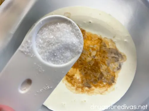 Sugar in a white measuring spoon being added to a bowl filled with heavy cream.