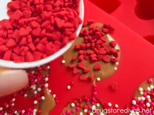 Heart-shaped sprinkles in a small bowl being poured on top of melted chocolate in a silicone mold.