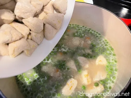 Biscuit pieces in a white bowl being poured into a Dutch oven with peas floating on top of a broth.