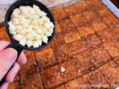 White chocolate chips in a measuring cup above a tray of toffee coated saltine crackers.