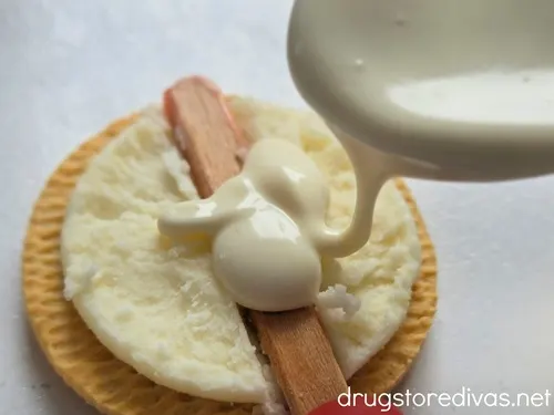A lollipop stick in a divot in the cream of a Golden Oreo cookie with a spoon pouring melted white chocolate on top.