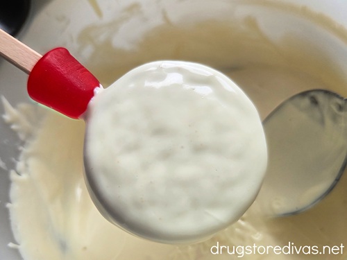 A dipped Oreo cookie with a red Dot candy on a stick above a bowl of melted white chocolate.