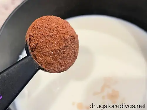 A measuring spoon filled with hot chocolate mix about to be poured into milk in a saucepan.