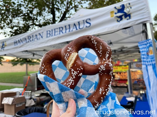 A hand holding a giant pretzel in blue and white checkered paper in front of a food tent at Nashville Oktoberfest.