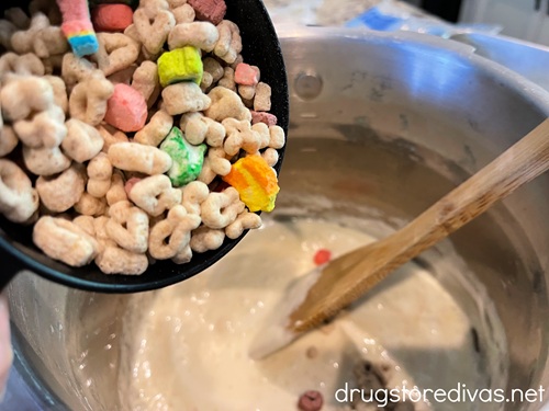 Lucky Charms cereal in a measuring cup above a stockpot of melted marshmallow that has a bamboo spoon in it.
