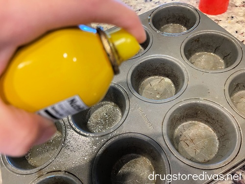 A hand holding a bottom of cooking spray that's spraying a muffin tin.