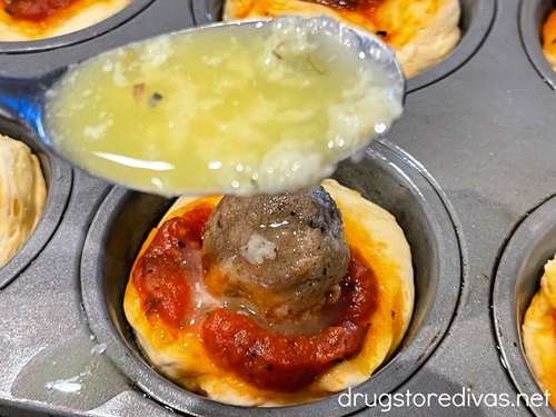 A spoonful of melted butter above to be poured on top of a meatball on top of marinara on a biscuit in a muffin tin.