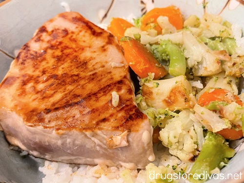 A cooked tuna steak next to cooked carrots, broccoli, and cauliflower over a bed of rice in a blue bowl.