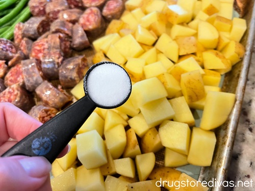 A measuring spoon with salt above green beans, cut sausage rounds, and cubed potato on a sheet pan.