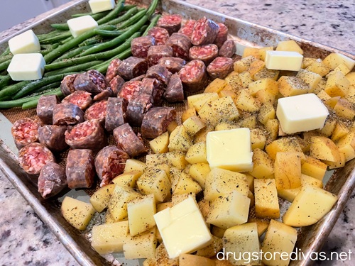Slices of butter on green beans, cut sausage rounds, and cubed potato on a sheet pan.