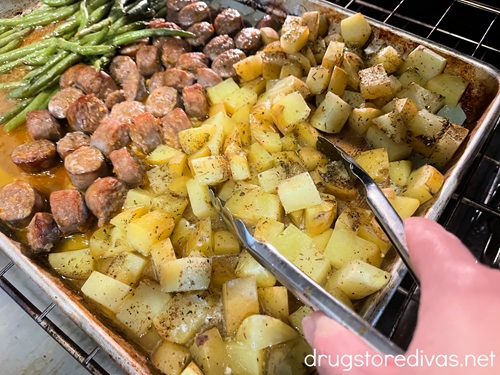 A hand holding a pair of tongs above green beans, cut sausage rounds, and cubed potato on a sheet pan.