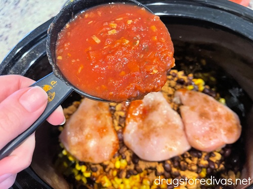 A hand holding a measuring cup filled with salsa above three pieces of seasoned chicken on top of corn and beans in a slow cooker.