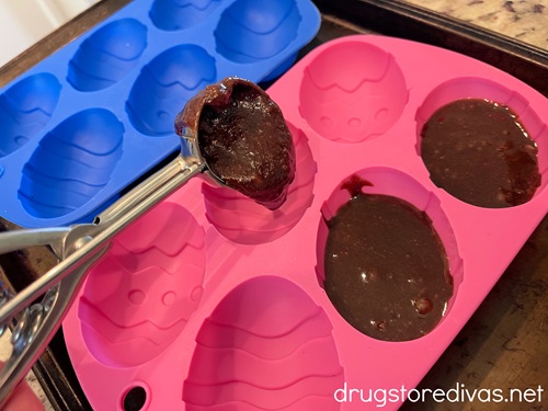 Brownie batter in a cookie scoop being poured into a pink silicone mold with egg shaped in it and a matching blue mold in the back.