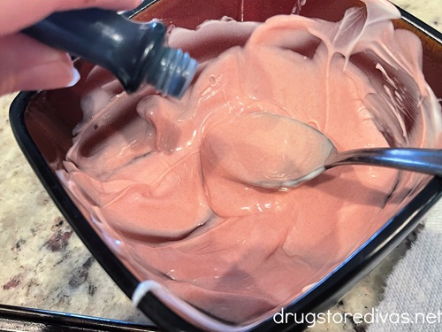 Pink melted chocolate in a bowl with a spoon to one side and a hand adding food coloring to the other.