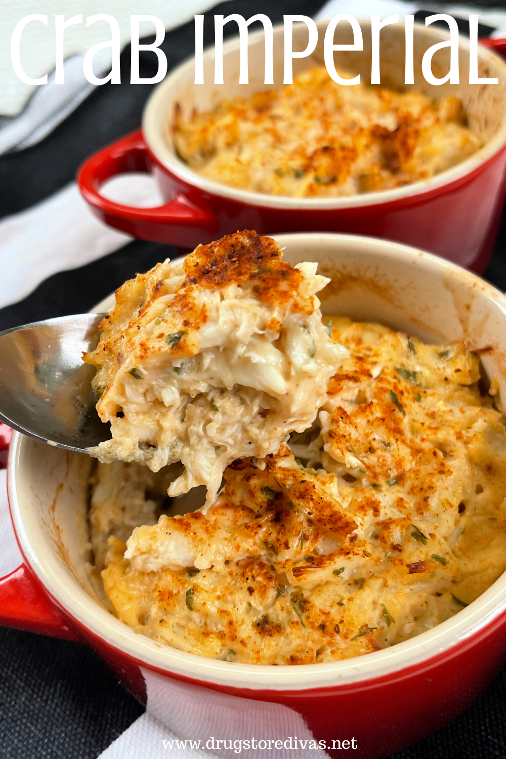 Two red, round cocottes filled with cooked crab meat, one with a spoon with some sticking out, on a black and white tablecloth with the words 