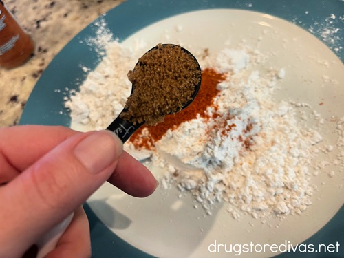 A hand holding a measuring spoon with seasoning in it above a plate with flour in it.