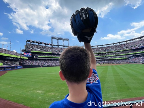 The back of the head of a little boy in a blue shirt who is holding up a baseball glove with his right hand at a baseball stadium.