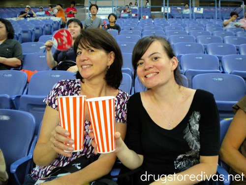 Two women holding orange and white striped cups in a Japanese baseball stadium.