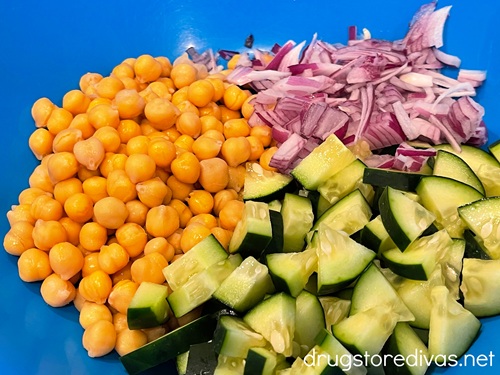 Cut cucumbers, sliced red onions, and chickpeas in a blue bowl.