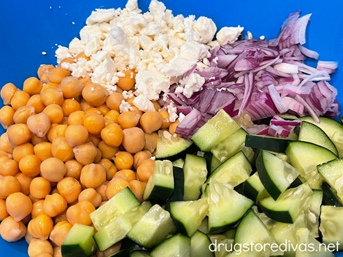 Cut cucumbers, sliced red onions, crumbled feta, and chickpeas in a blue bowl.