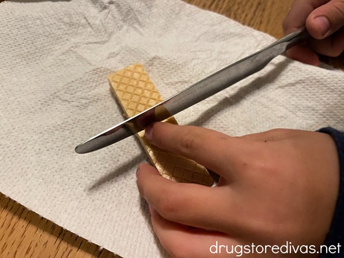 A child's hand holding a butter knife and cutting a vanilla wafer cookie.