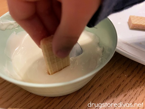 A child's hand dipping a piece of wafer cookie into melted white chocolate.
