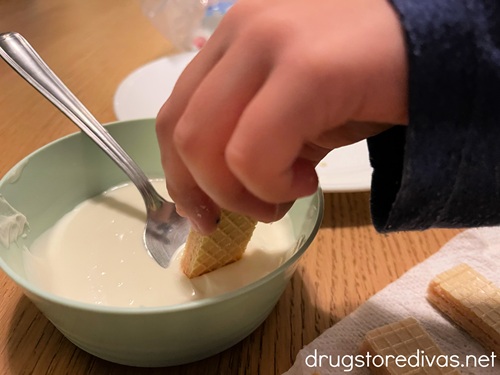 A child's hand dipping a piece of wafer cookie into melted white chocolate.