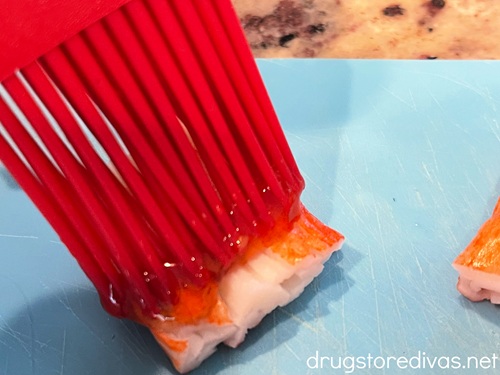 A red pastry brush brushing sauce on a piece of imitation crab meat that's on a blue cutting board.