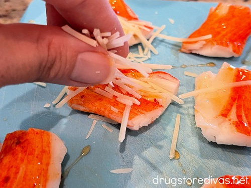A hand sprinkling fancy shredded Parmesan cheese on top of smashed imitation crab pieces on a white cutting board.