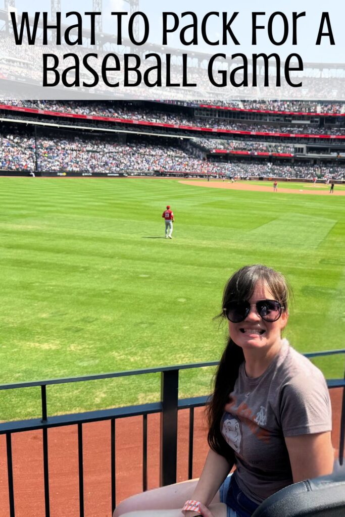 A woman sitting in the first row at a baseball stadium with the field and a player on it behind her and the words "What To Pack For A Baseball Game" digitally written above her.