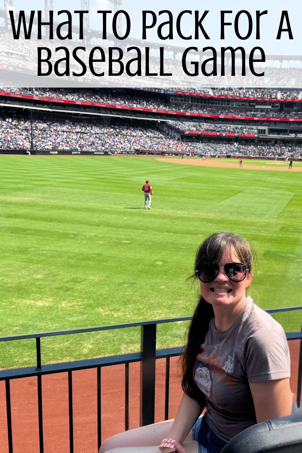 A woman sitting in the first row at a baseball stadium with the field and a player on it behind her and the words 