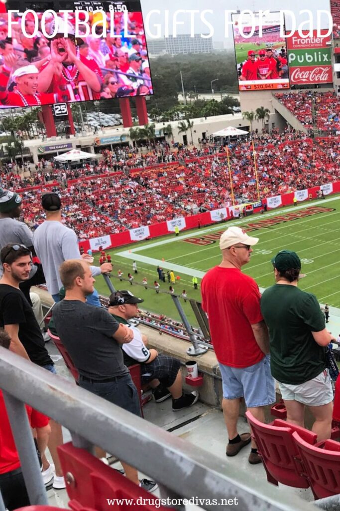 Men in the Tampa Bay Buccaneers football stadium watching a game with the words "Football Gifts For Dad" digitally written on top.