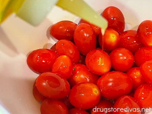 A bottle with olive oil in it being poured on top of cherry tomatoes in a white bowl.