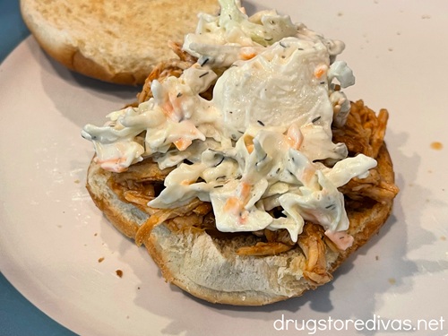 Coleslaw and barbecue pulled chicken on the bottom half of a hamburger bun with the top nearby.