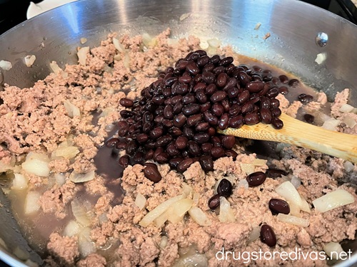 Cooked ground turkey, diced onions, and black beans being stirred with a bamboo spoon in a silver pan.