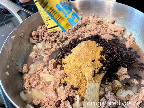 A packet of taco seasoning being poured on ground turkey, black beans, and diced onions in a silver plan.