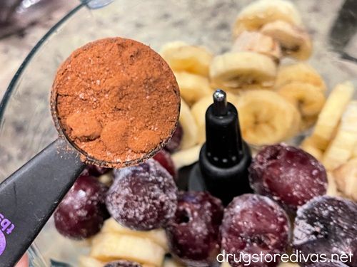 Cocoa powder in a measuring spoon above cherries and banana slices in a blender.