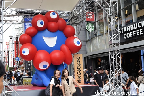Two women posing in front of a red and blue figure, the mascot for the Osaka Expo, in Dotonbori Japan.