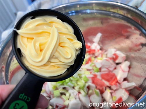 A measuring cup with mayo in it above imitation crab and celery pieces in a silver bowl.