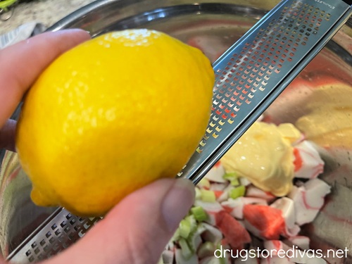 A hand holding a lemon on a microplane grater above imitation crab and celery pieces in a silver bowl.