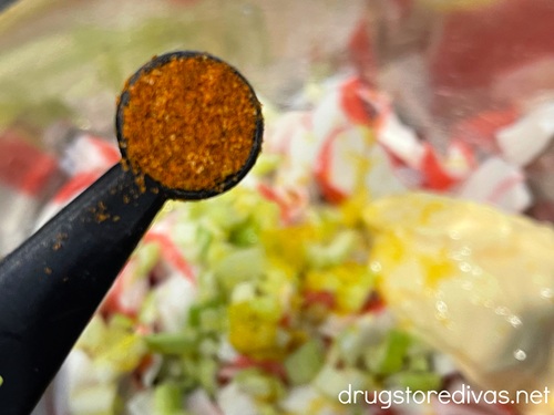 A measuring spoon with red seasoning in it above imitation crab and celery pieces in a silver bowl.