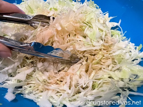 Tongs tossing shredded cabbage in a blue bowl.