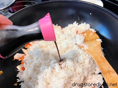 Soy sauce in a bottle being poured into a wok with imitation crab pieces, white rice, and a bamboo spoon in it.