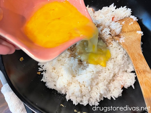 Scrambled eggs in a pink bowl being poured into a wok with imitation crab pieces, white rice, and a bamboo spoon in it.