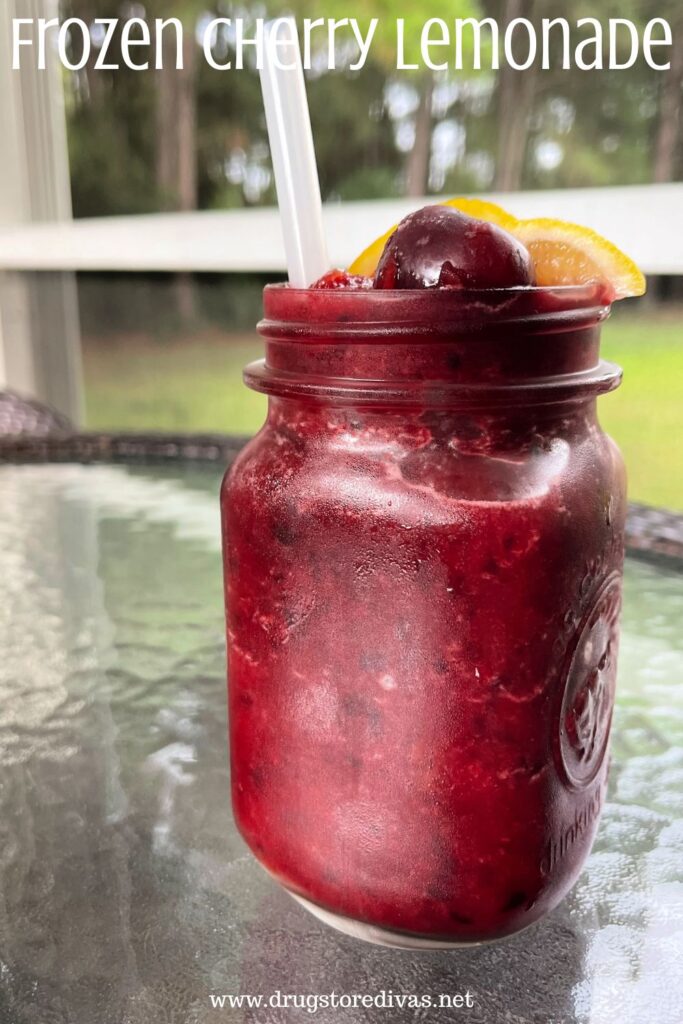 A maroon frozen drink in a mason jar with a straw, cherry, and lemon wedges coming out of the top and the words "Frozen Cherry Lemonade" digitally written above it.