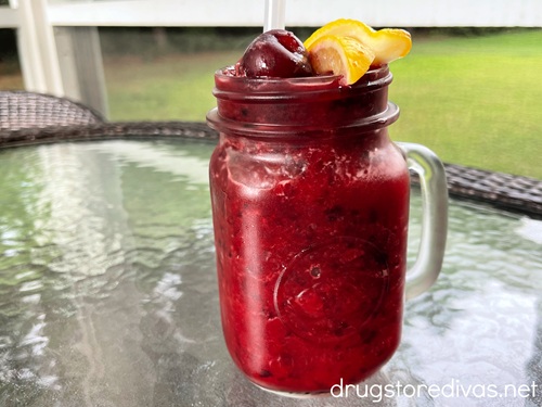 A maroon beverage in a mason jar with a handle with a straw, a frozen cherry, and lemon slices on top.