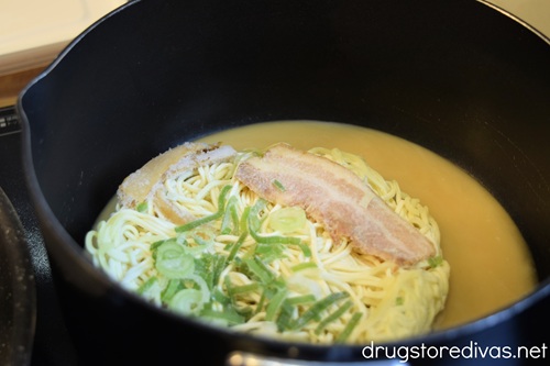 Frozen ramen noodles, green onion, and bacon with broth in a black stockpot on a stove.